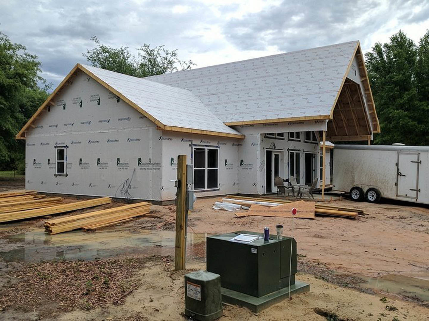 Two-story house under construction with exposed lumber framing, partially finished roof, and a wheeled trailer parked on dirt ground; nearby trees and cloudy sky in background.