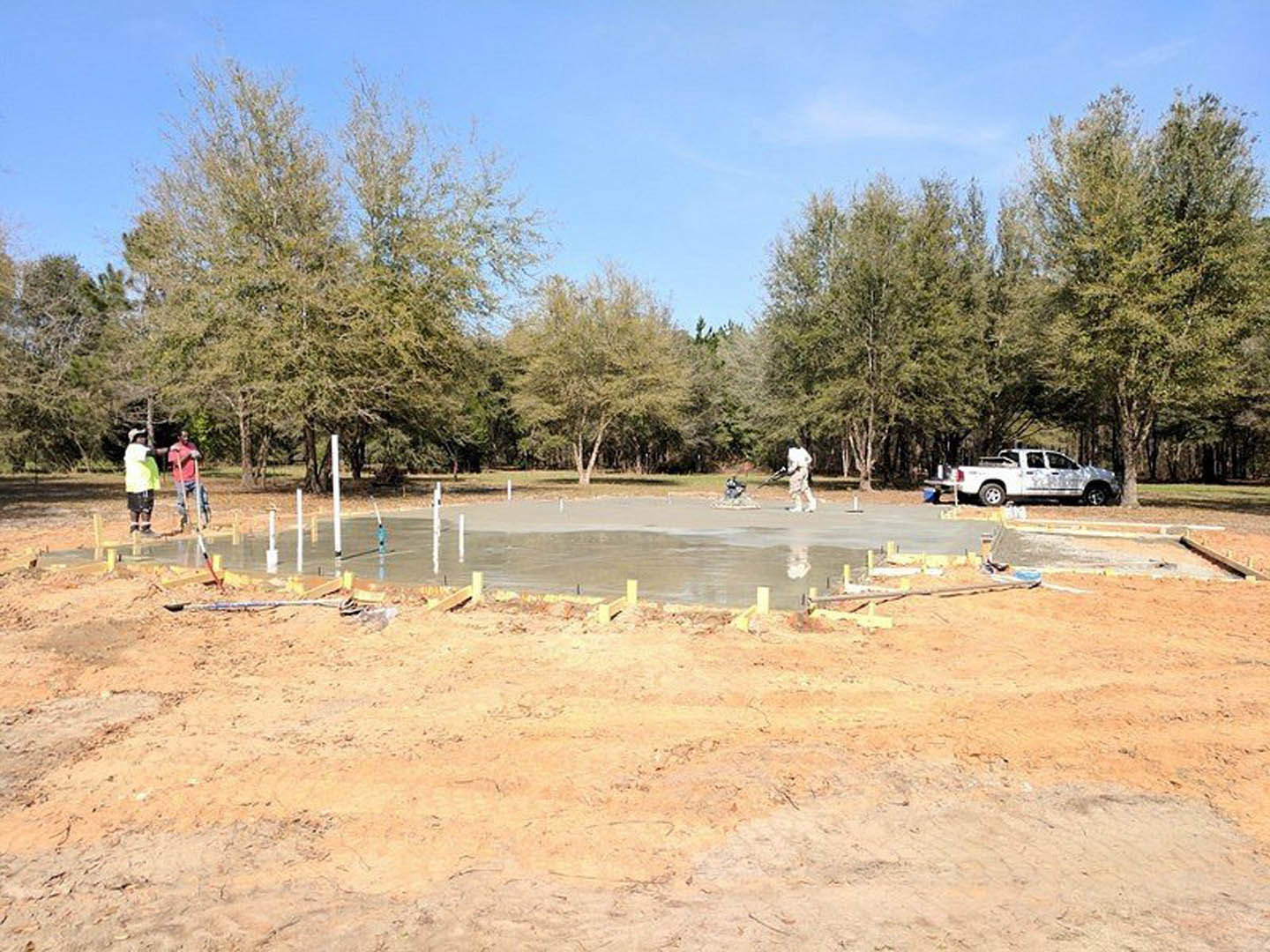 Framed custom home under construction with exposed wooden beams, several workers on site, white truck parked nearby, dirt ground, scattered tools, and trees in the background under