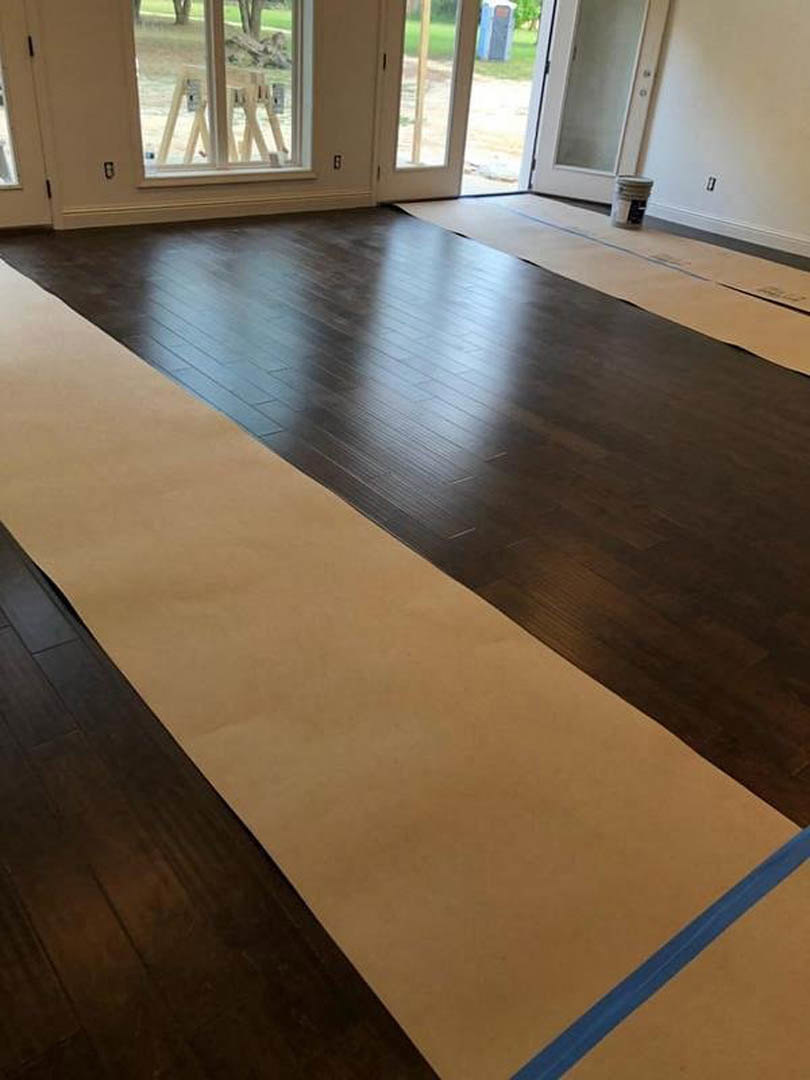 Spacious room with light wood plank flooring, oversized wooden door, large window framed in wood, and cardboard sheet with blue line placed on the floor