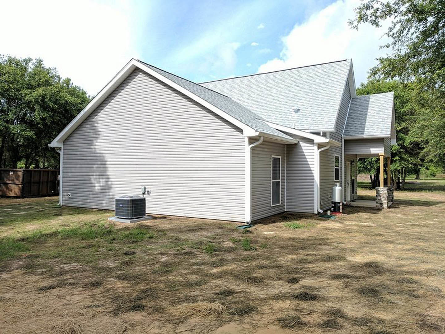 Two-story house with gray siding, gabled roof, and white trim, surrounded by green lawn and mature trees under a partly cloudy sky