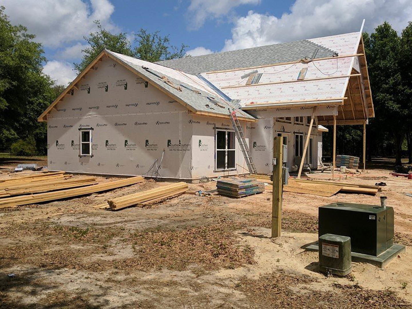Framed house under construction with exposed wood planks, ladder leaning against exterior, metal waste container and stacked boxes on dirt ground, cloudy sky and trees in