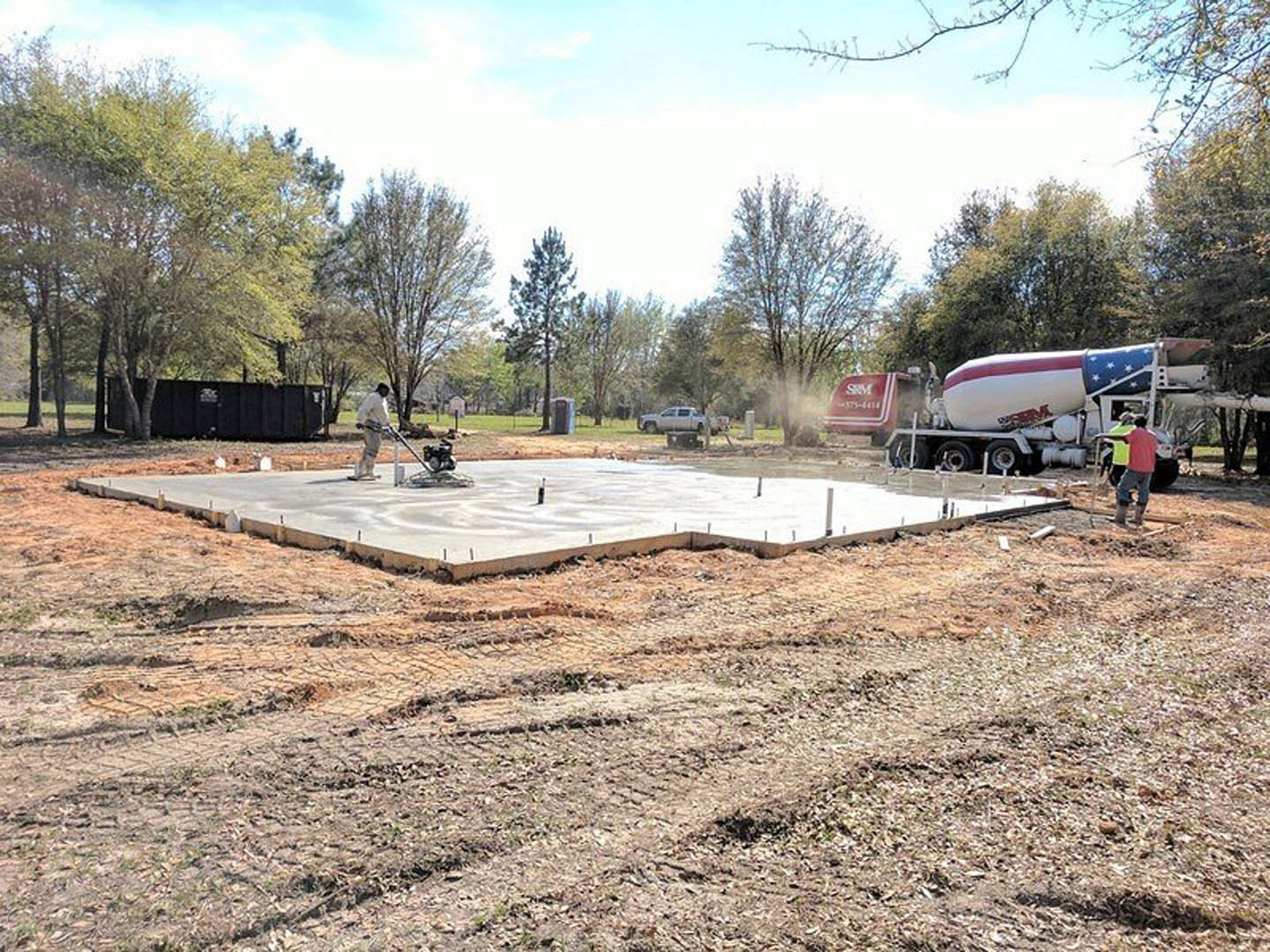 Man in pink shirt standing on newly poured concrete slab next to cement mixer, with construction materials and trees visible in background
