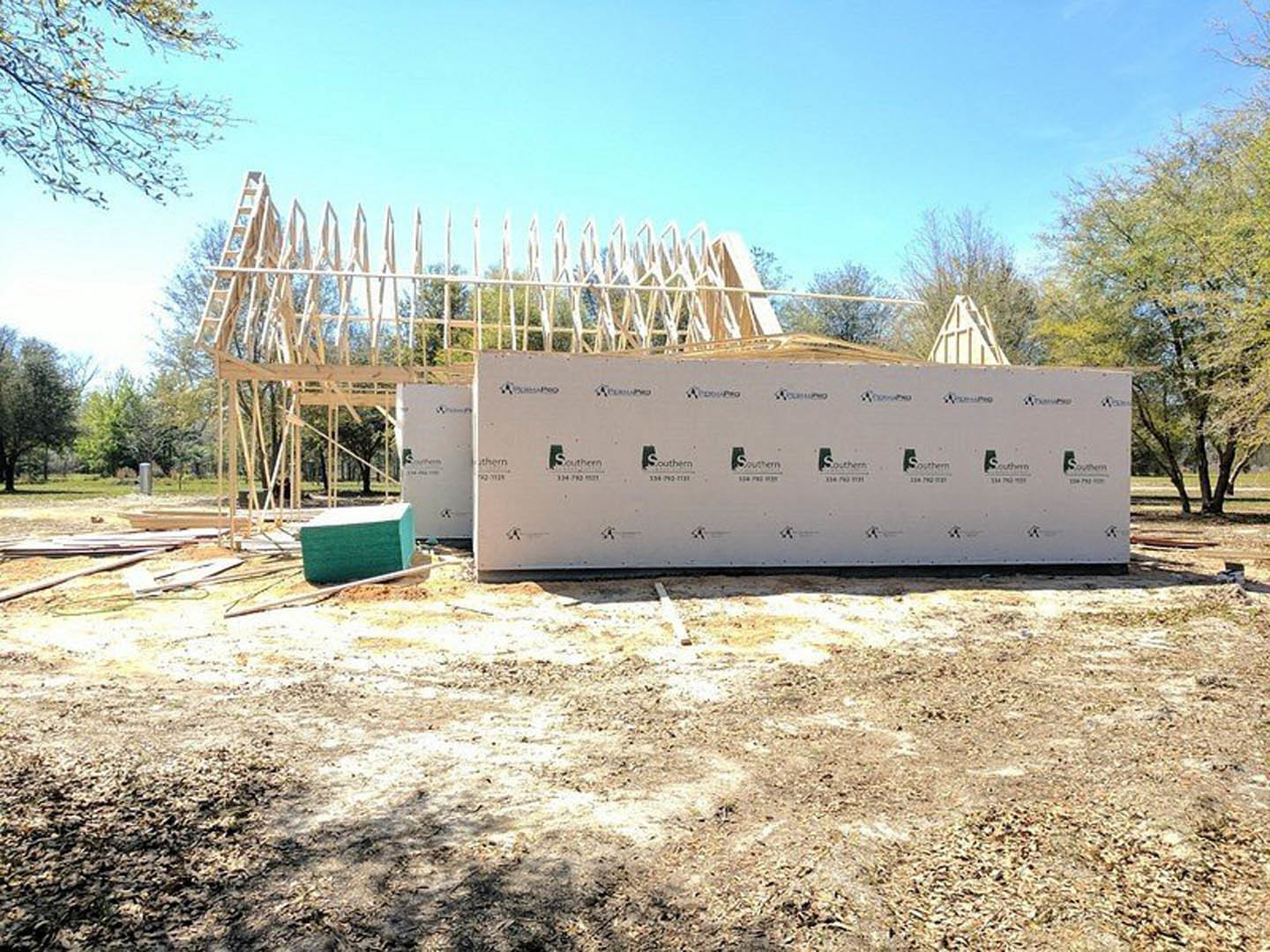 Wood-framed custom home under construction with exposed beams, white temporary wall in dirt, blue sky overhead, and trees in background