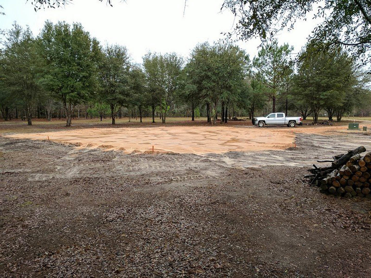 White pickup truck parked on a dirt lot surrounded by scattered logs and mature trees, grassy patches visible in the background.
