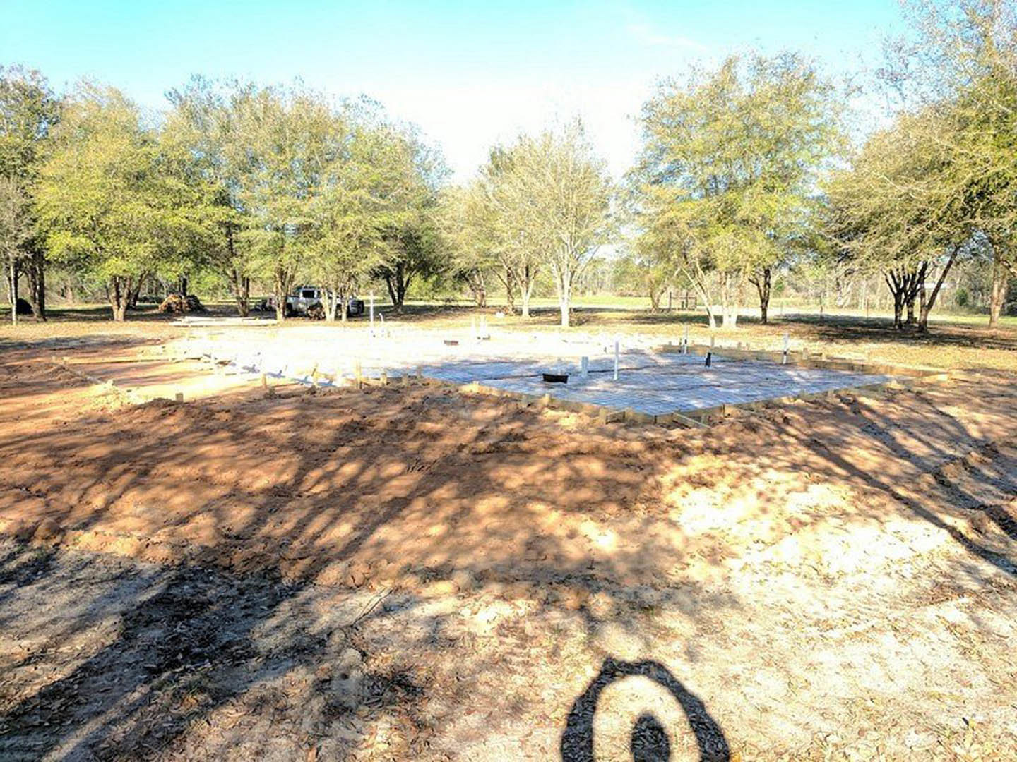 Dirt construction site bordered by mature trees, wooden deck partially built, open sky above, grassy landscape in background