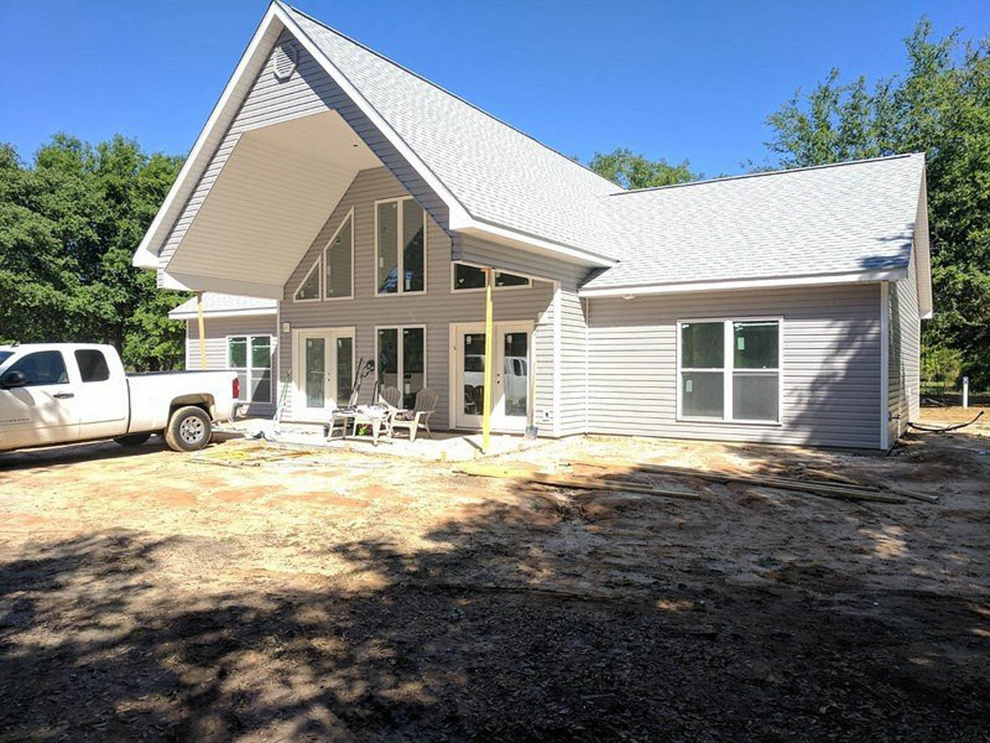 Two-story house with gray roof and covered porch, white pickup truck parked in dirt yard, open truck door, folding chairs near yellow pole, large windows visible on facade