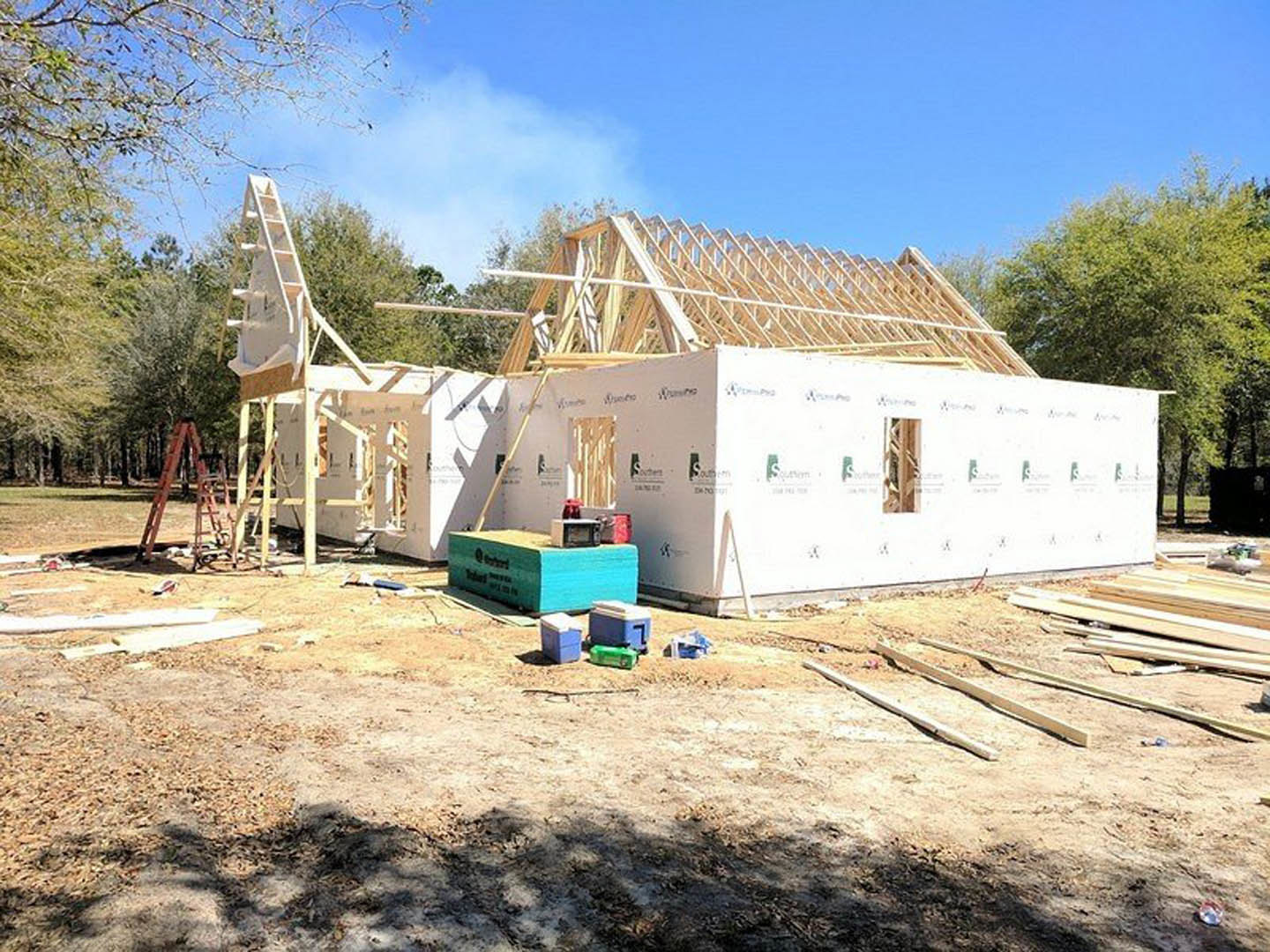 Wood-framed house under construction with exposed wooden roof, two ladders leaning against exterior wall, white box with logo, blue cooler, and several coolers grouped on bare