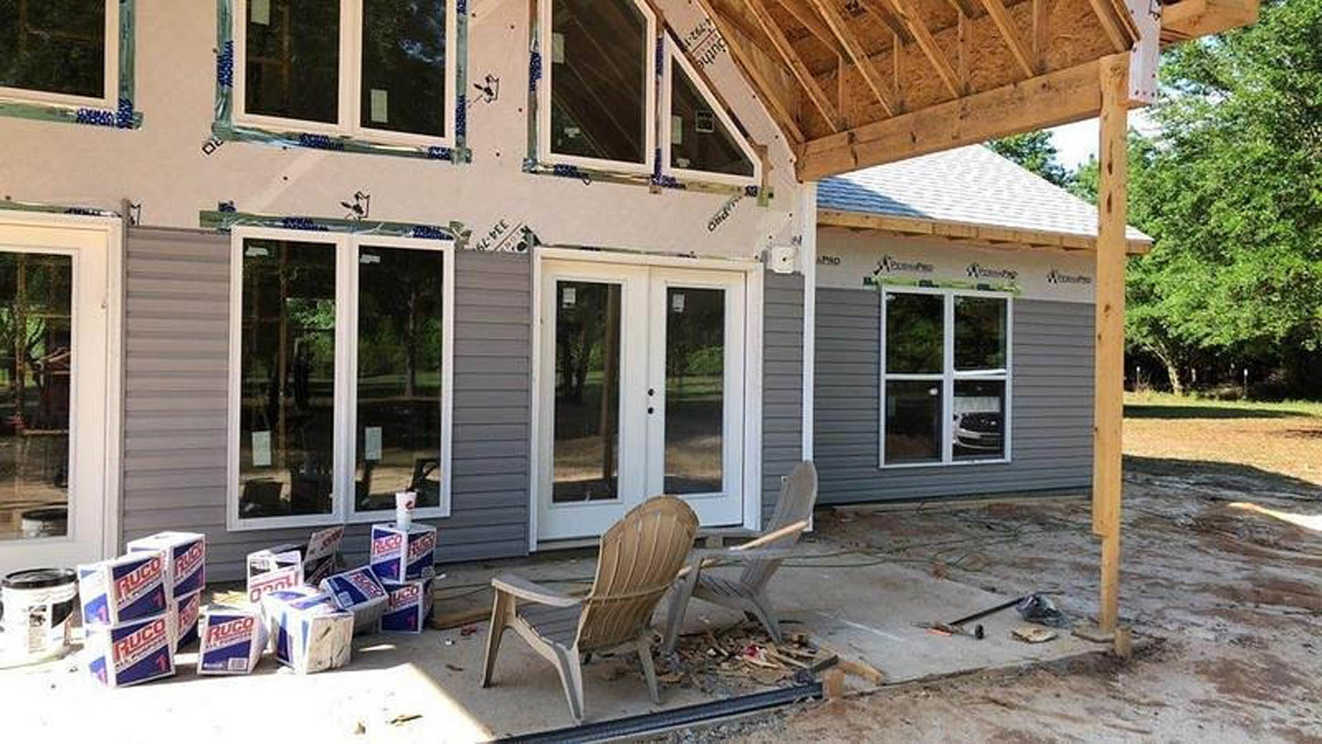 White-framed glass double doors and windows on a partially constructed home, wooden chair with a stick and boxes on the shaded porch, tree branches overhead