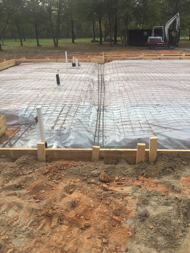 Concrete foundation under construction with wire mesh reinforcement, wooden beams, dirt and rocks scattered on site, white tarp covering part of the area, backhoe parked with door