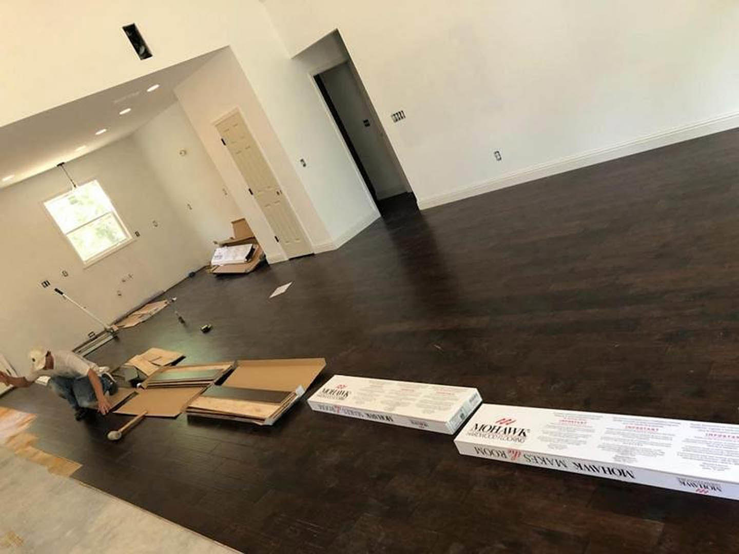 Spacious room with polished hardwood flooring, several cardboard boxes scattered across the floor, white plaster walls, and natural light streaming through a window