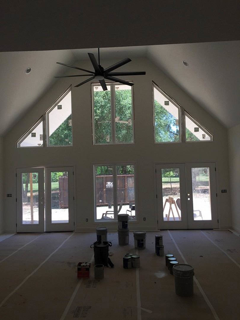 White ceiling fan with light fixture above a room with beige walls, several paint cans and buckets scattered on the light-colored floor, window providing daylight.