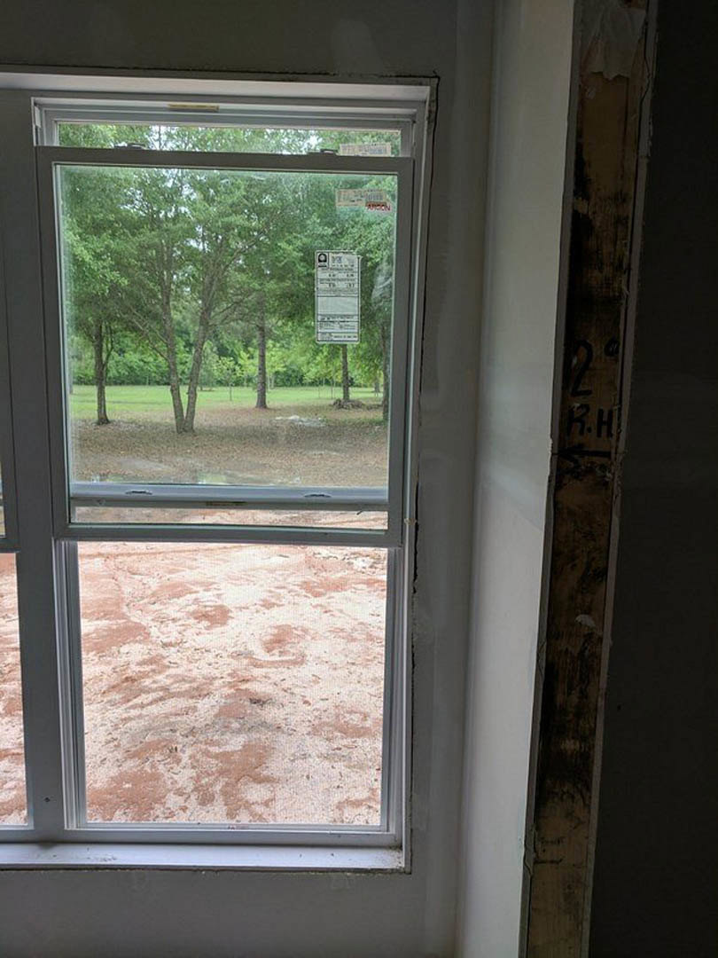 Large window framed in white trim, overlooking leafy green trees; sunlight streams into a neutral-toned room with sandy textured flooring, a black-lettered sign displayed on the