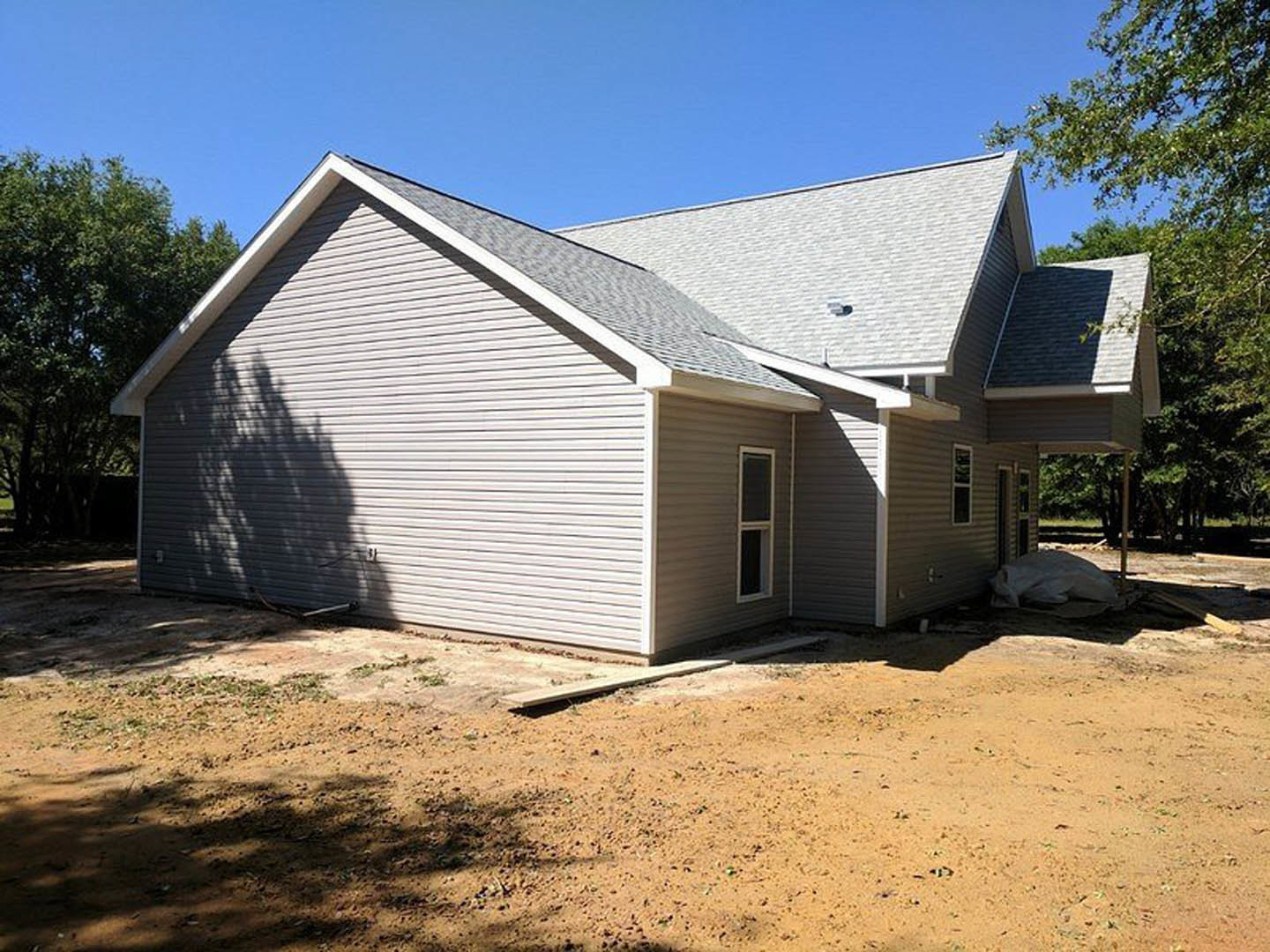 Partially built home with exposed wooden framing, shingled roof, dirt lot, and mature trees in the background