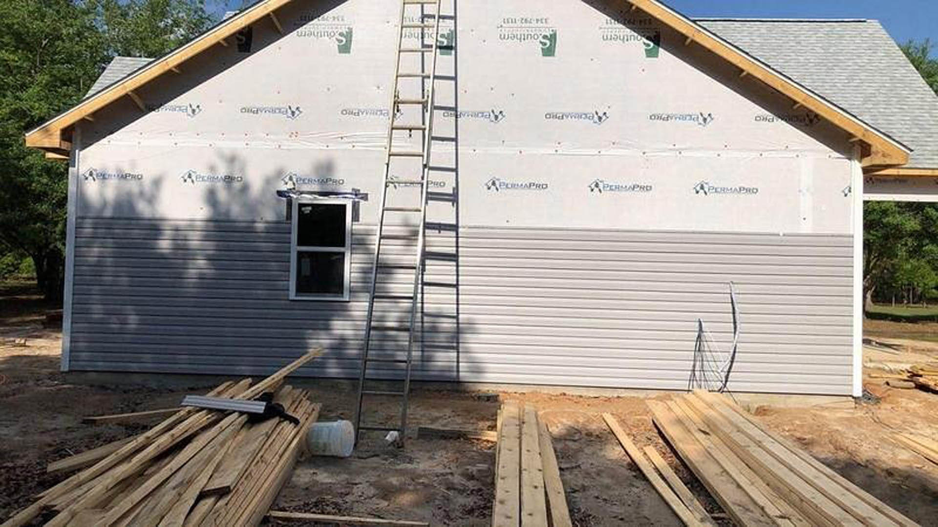 White siding house with black-framed window, wood planks stacked beside exterior wall, metal ladder leaning against building, leafy tree in background