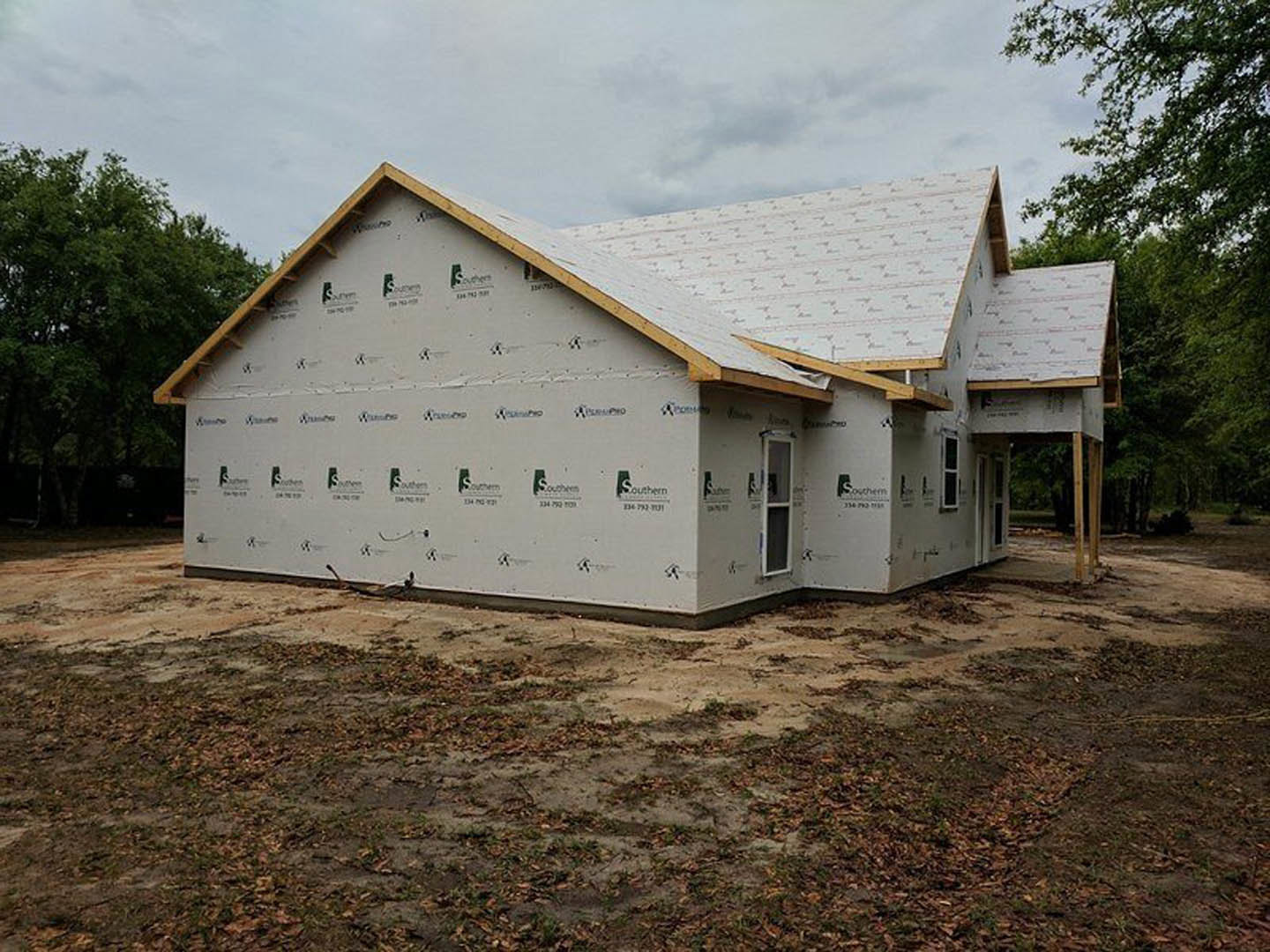 Partially built house with exposed framing, completed roof, large windows, and dry leaves scattered across dirt yard; trees visible in background under cloudy sky