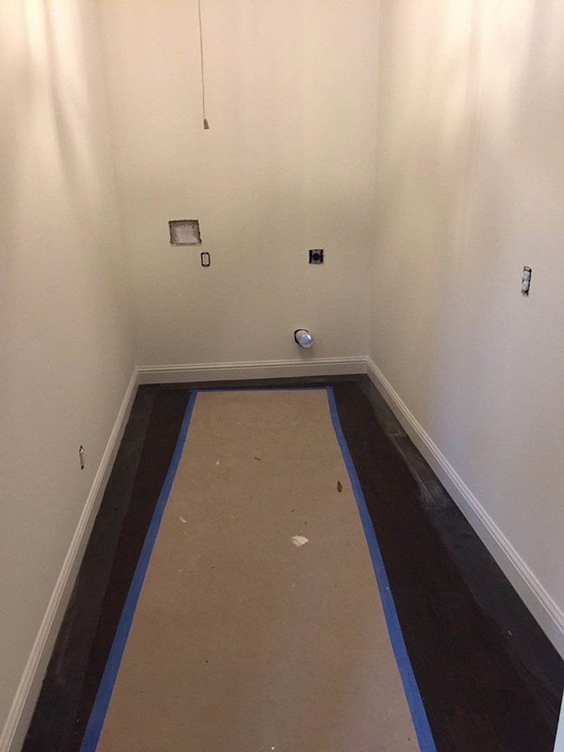Hallway with light-colored carpet featuring blue tape, white plaster walls, ceiling-mounted light fixture, and black-bordered square object near the entrance.