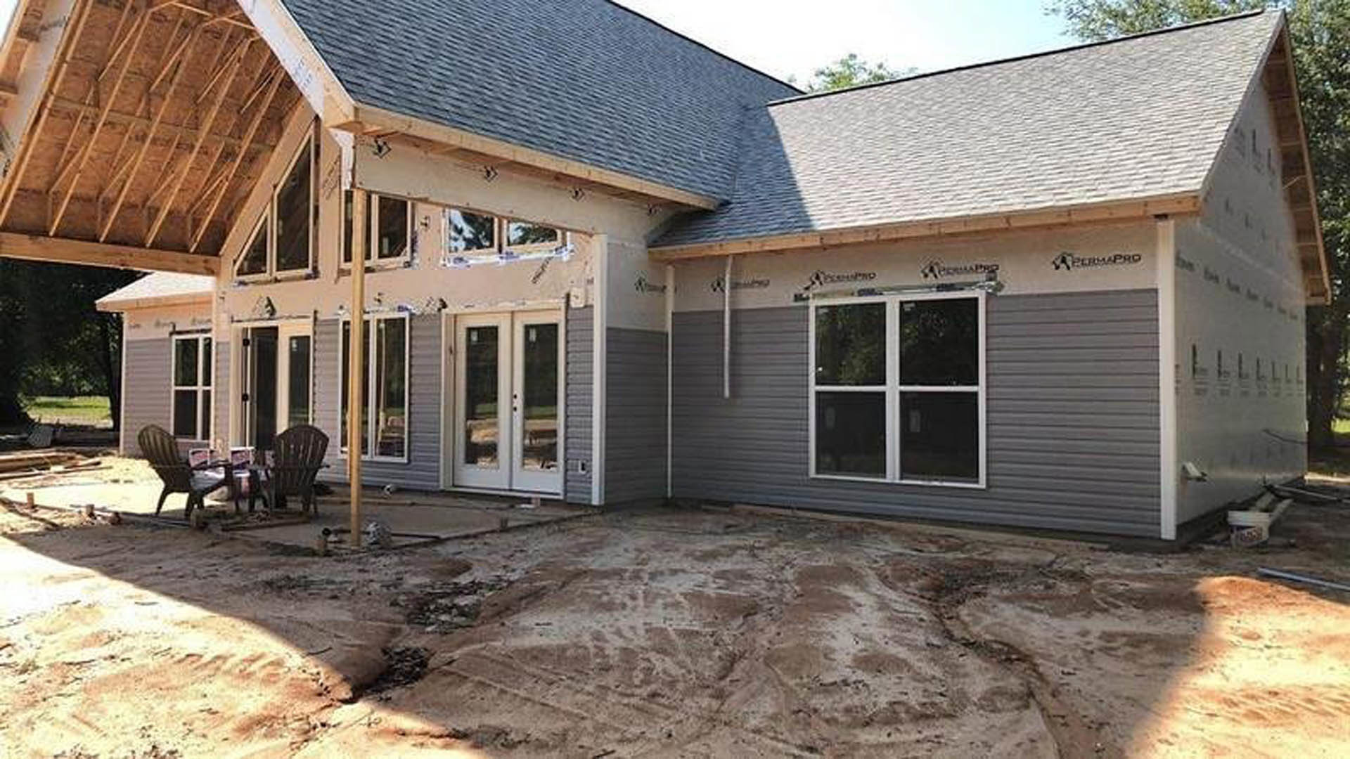Partially built house with exposed siding, white-framed windows, dirt yard, and two chairs placed near the entrance.