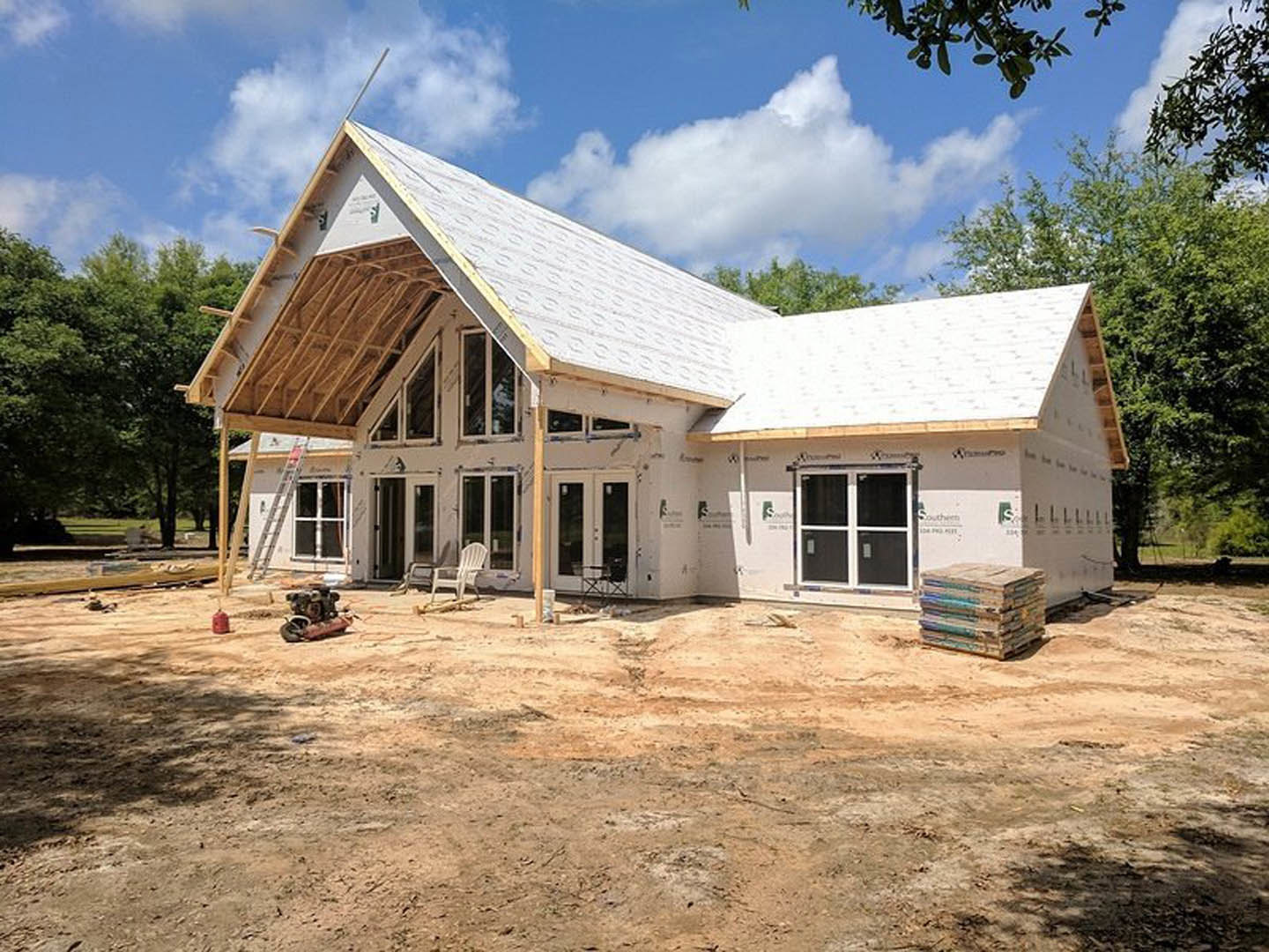 Wood-framed house under construction with exposed roof trusses, surrounded by mature trees and cloudy sky in background