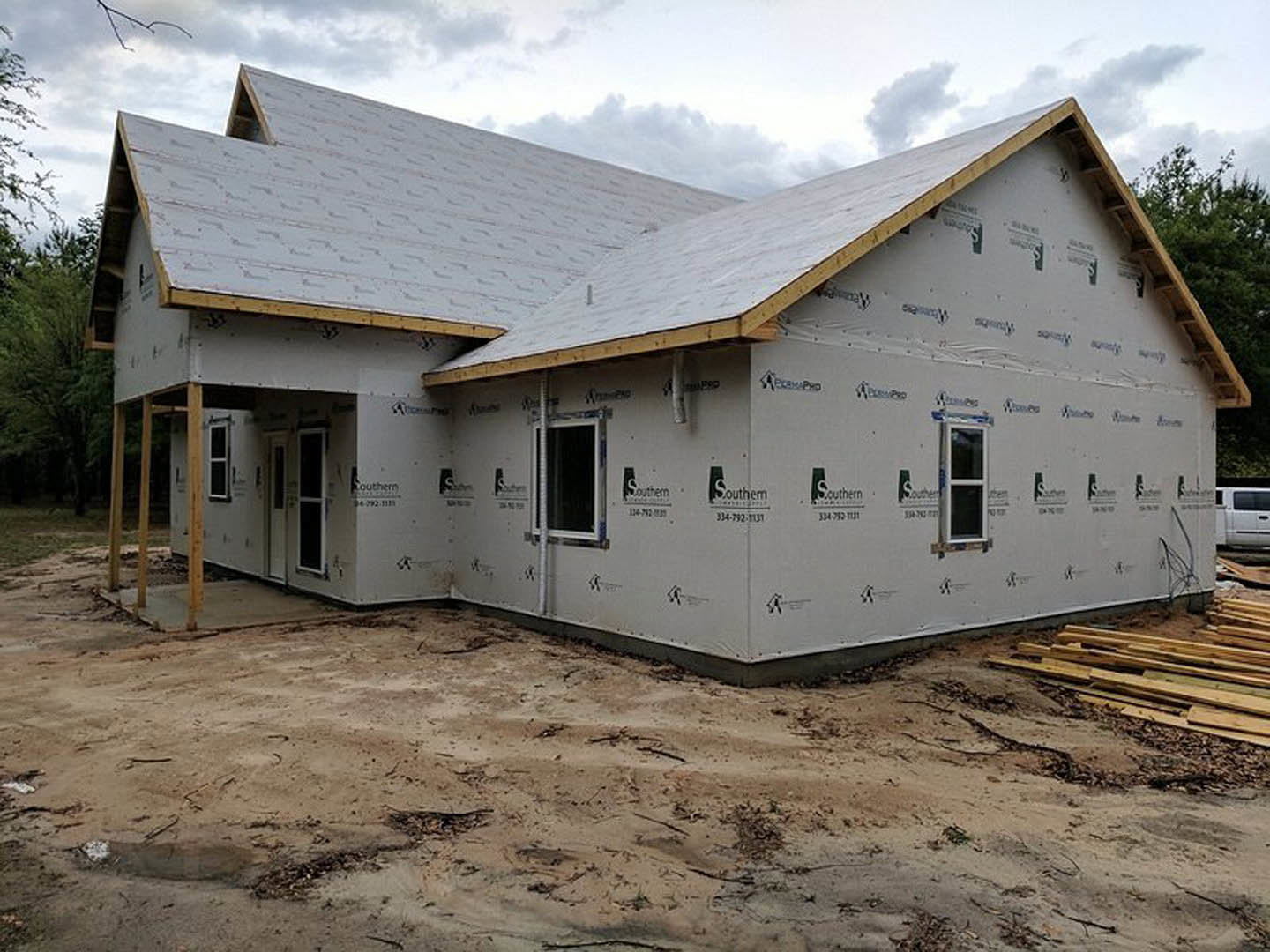 Partially built house with exposed wooden framing, roof installed, white van parked nearby, pile of lumber on dirt ground, white exterior wall with window featuring white trim