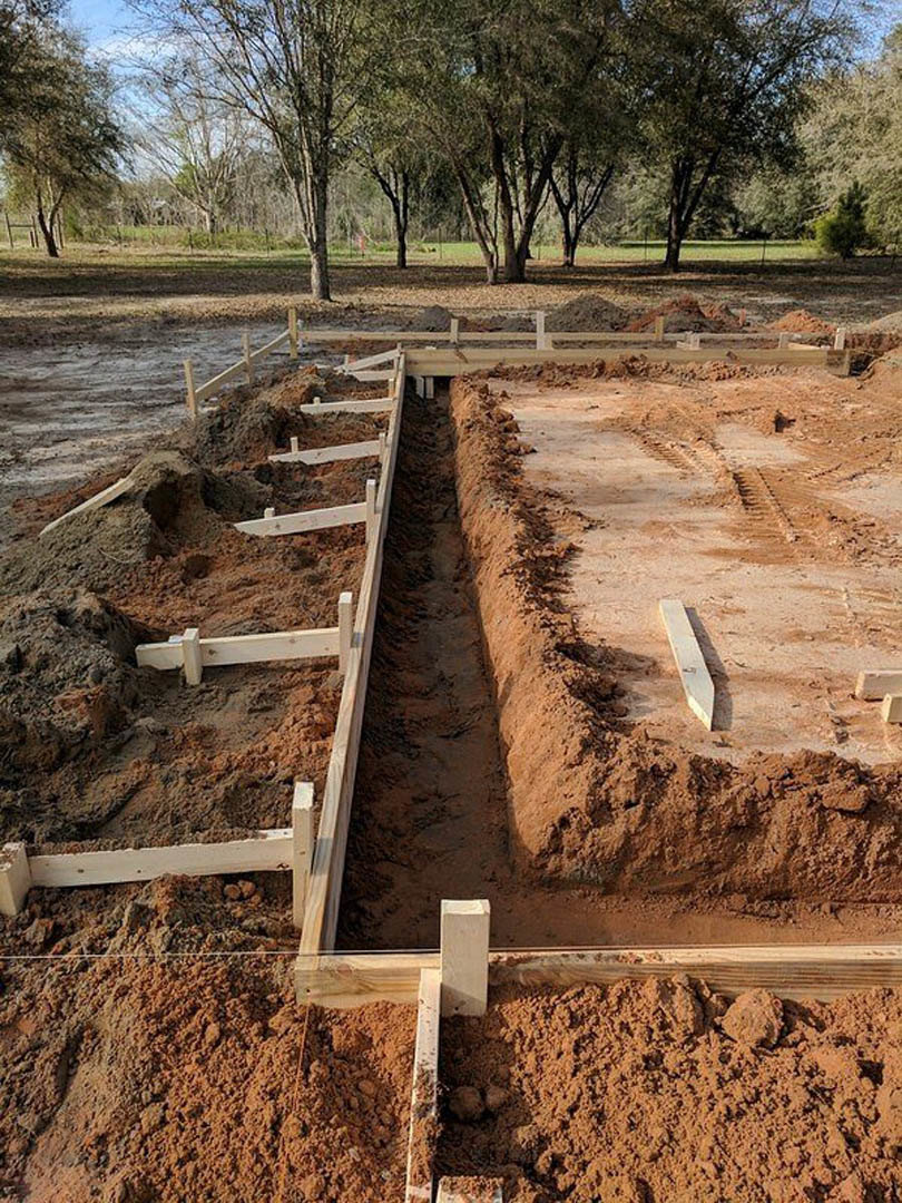 Wooden foundation forms set in dirt at a construction site, with a trench and tire tracks visible in the soil, surrounded by trees and open sky.