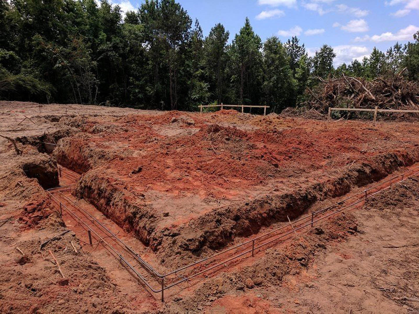 Dirt ground with scattered metal bars, wooden fence, and trees against a blue sky in the background