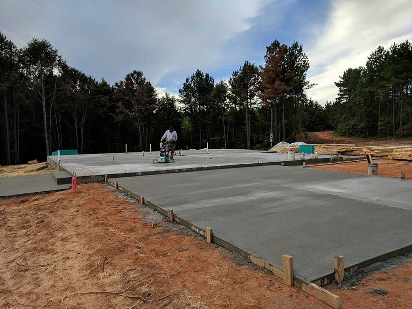 Man standing on unfinished concrete slab foundation surrounded by wood posts, with trees and cloudy sky in background