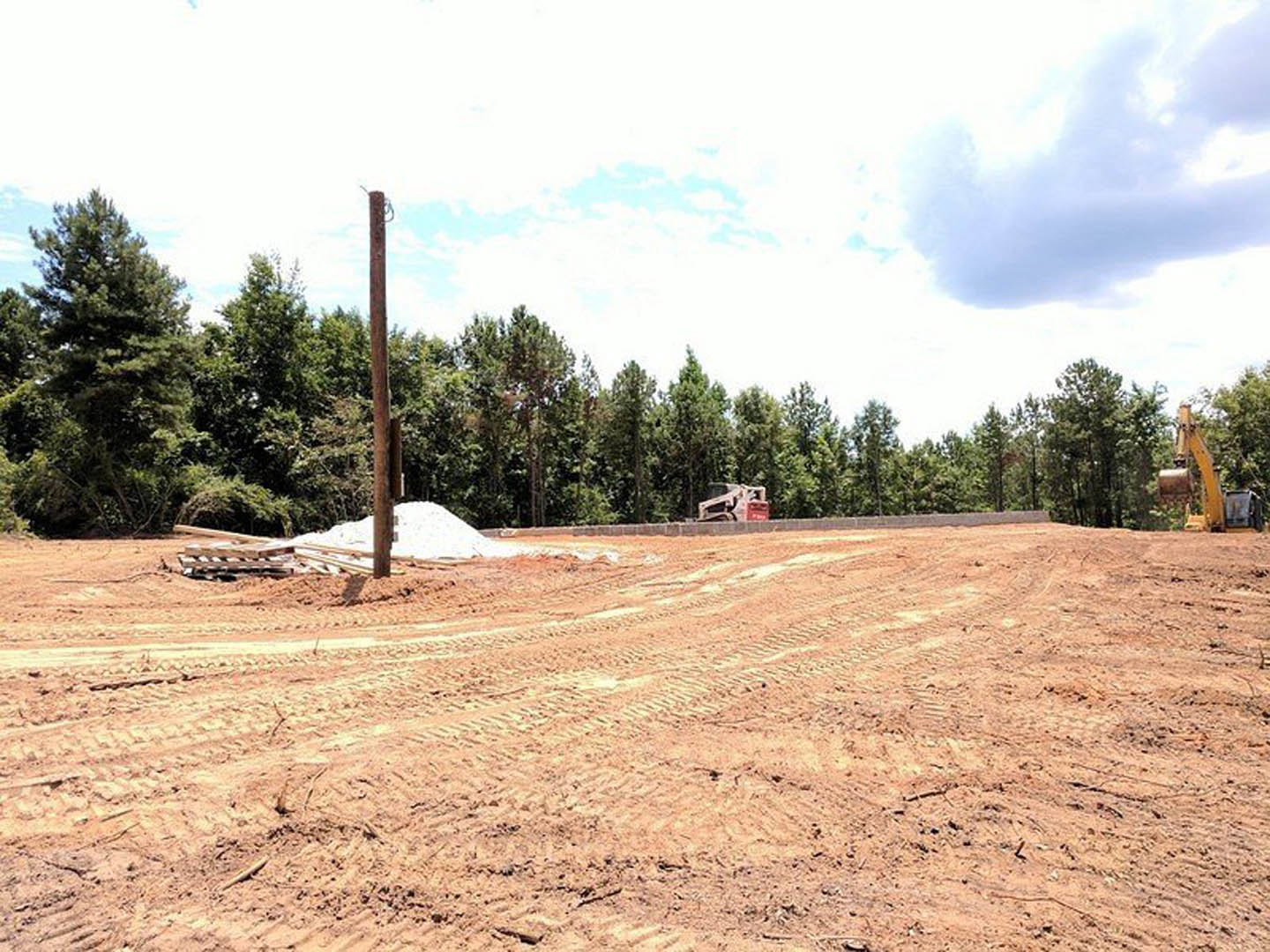 Dirt field with tire tracks and a large pile of soil, bordered by trees under a partly cloudy blue sky