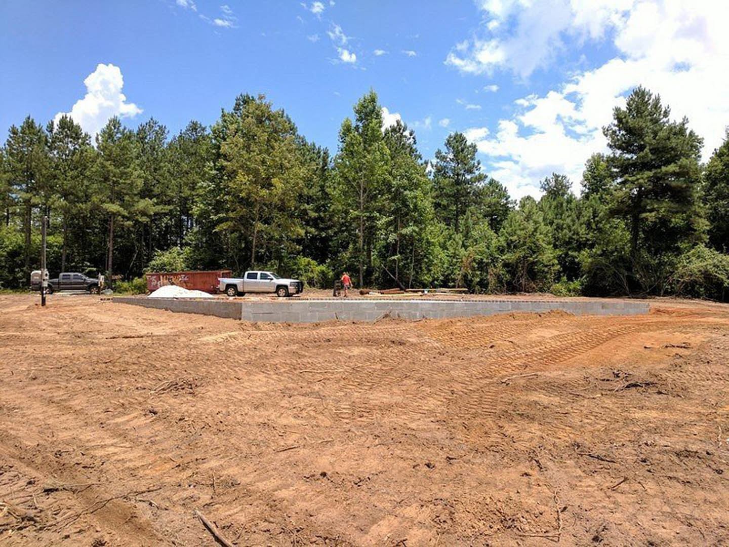 White pickup truck parked on a dirt field near a concrete wall, man standing in front, surrounded by trees under a blue sky with scattered clouds.