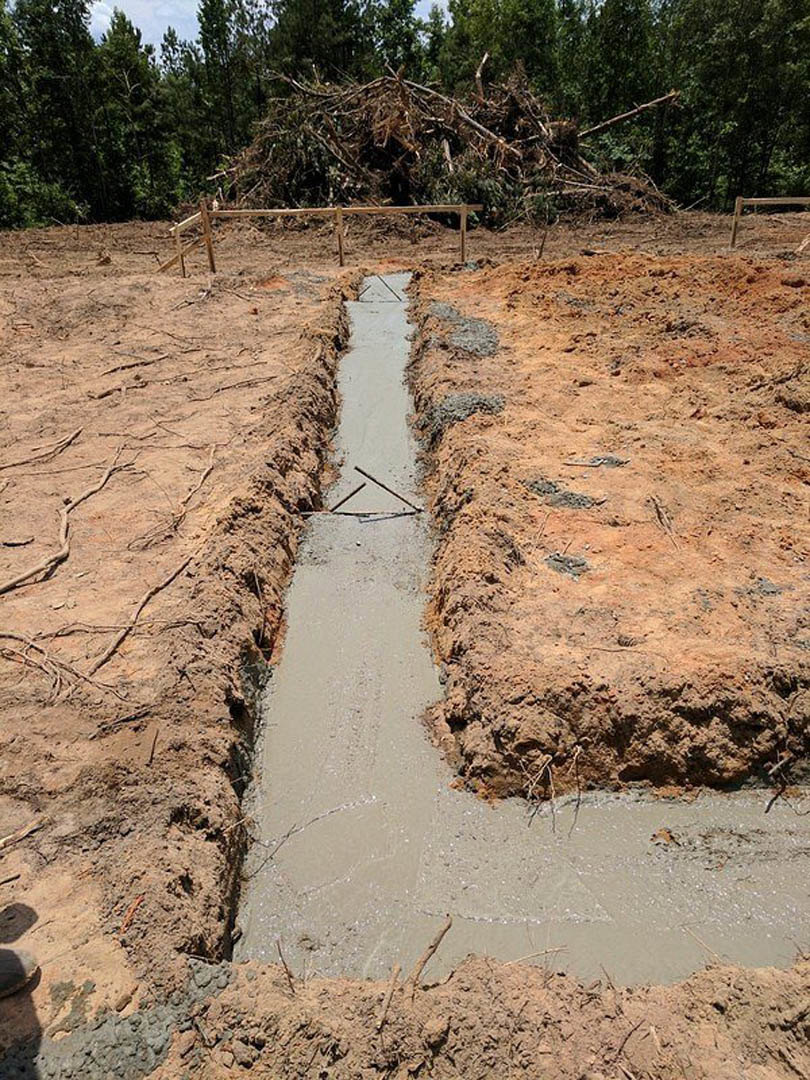 Concrete trench running through a dirt field, bordered by a wooden fence and scattered tree branches, with a white rectangular object and brown leaves nearby.