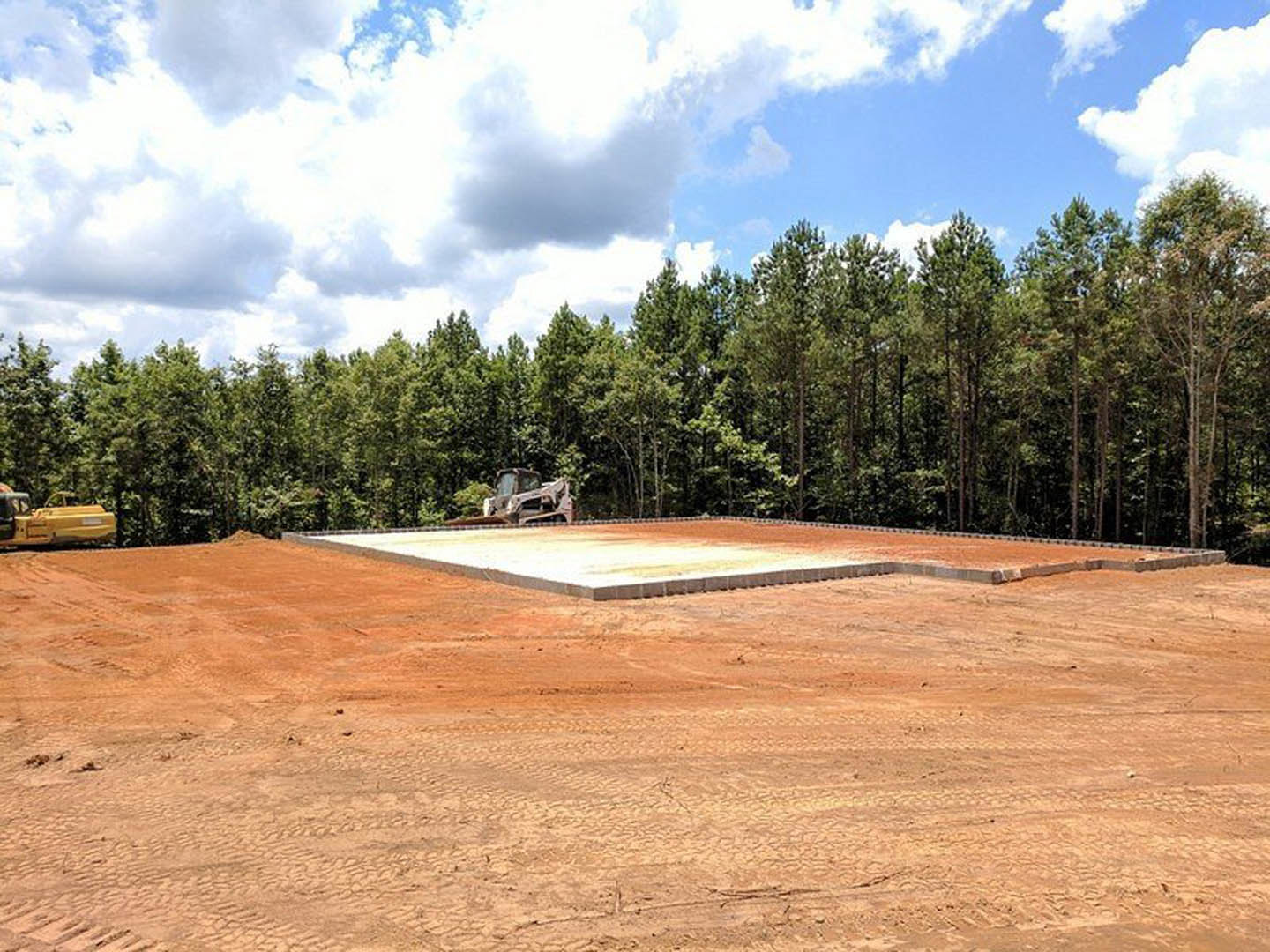 Square concrete foundation set in a large dirt clearing, surrounded by dense trees under a blue sky with scattered clouds; yellow construction vehicle and worker visible near the