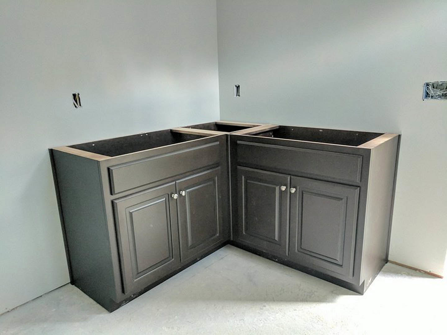 White shaker-style kitchen cabinets and drawers in a corner, light-colored walls, visible electrical box with white wires, wood flooring