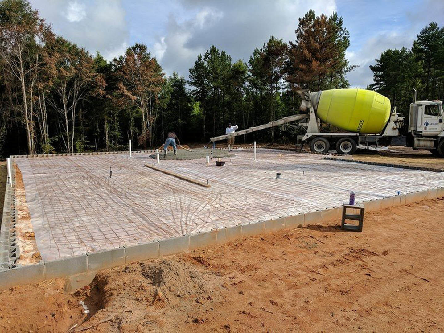 Cement mixer on trailer at construction site with workers, exposed soil, scattered tools, and trees under cloudy sky