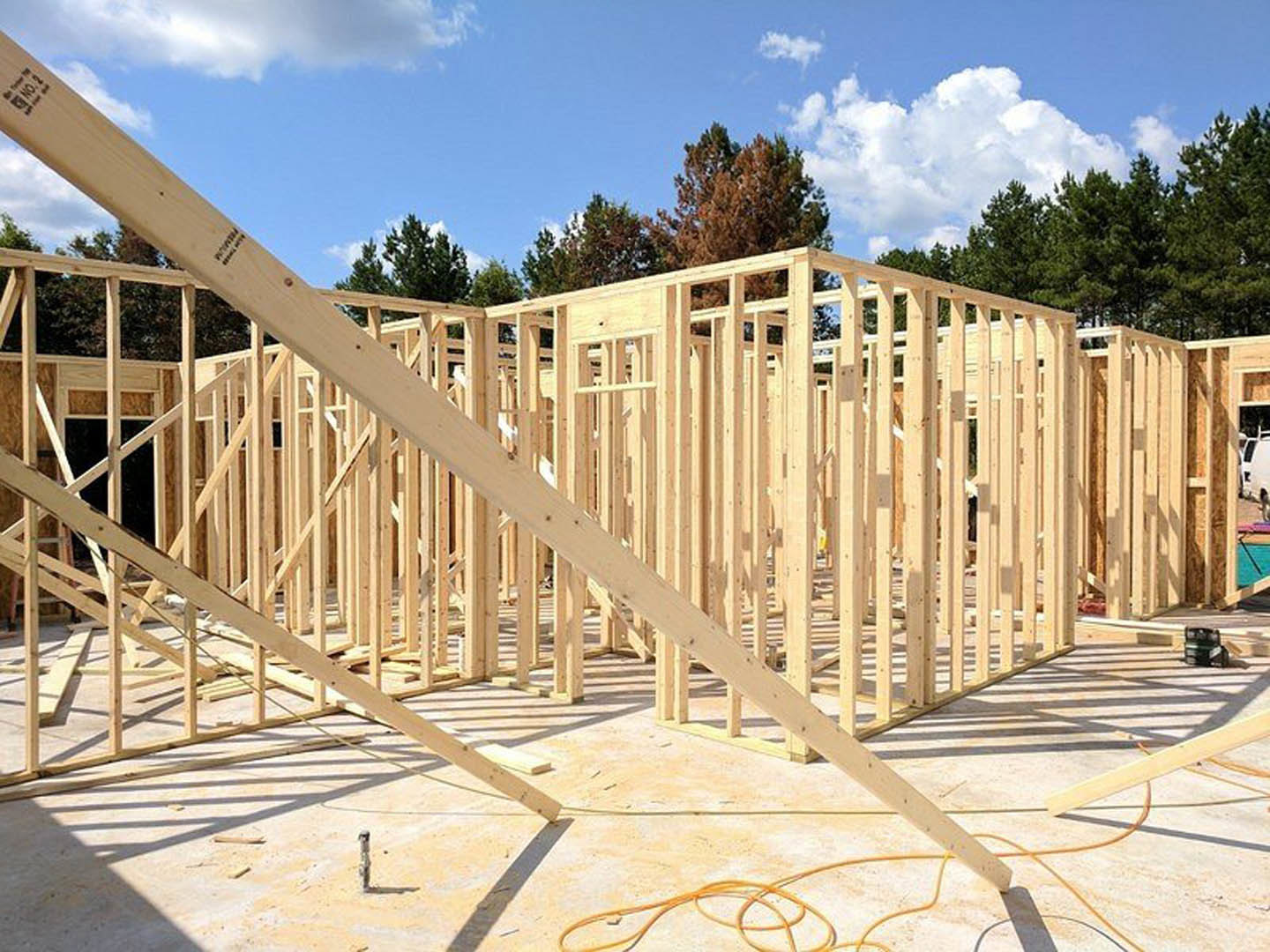 Wood-framed house under construction with exposed beams and plywood, set against a blue sky with scattered clouds and trees in the background
