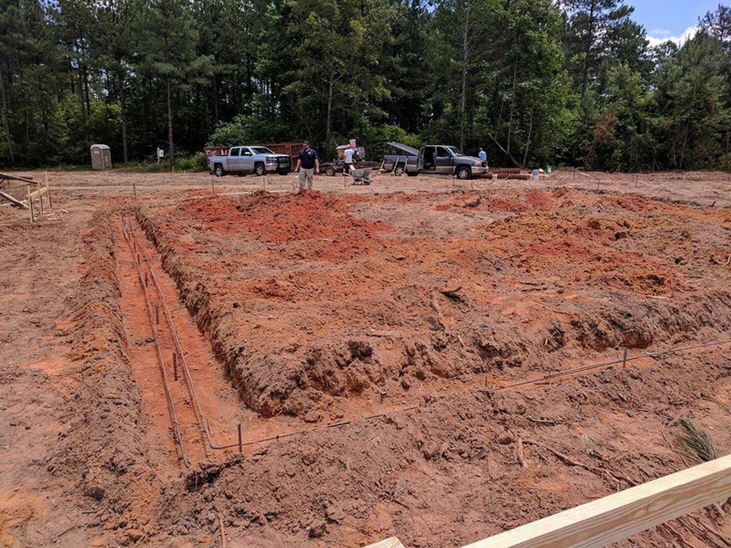 Construction site with several men standing on bare dirt, white pickup truck parked nearby with door open, portable toilet visible, trees and sky in background
