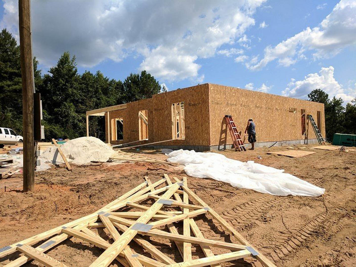 Wood-framed house under construction with exposed beams, ladder leaning against structure, pile of lumber on dirt, white sheet spread nearby, man walking past, white pickup truck