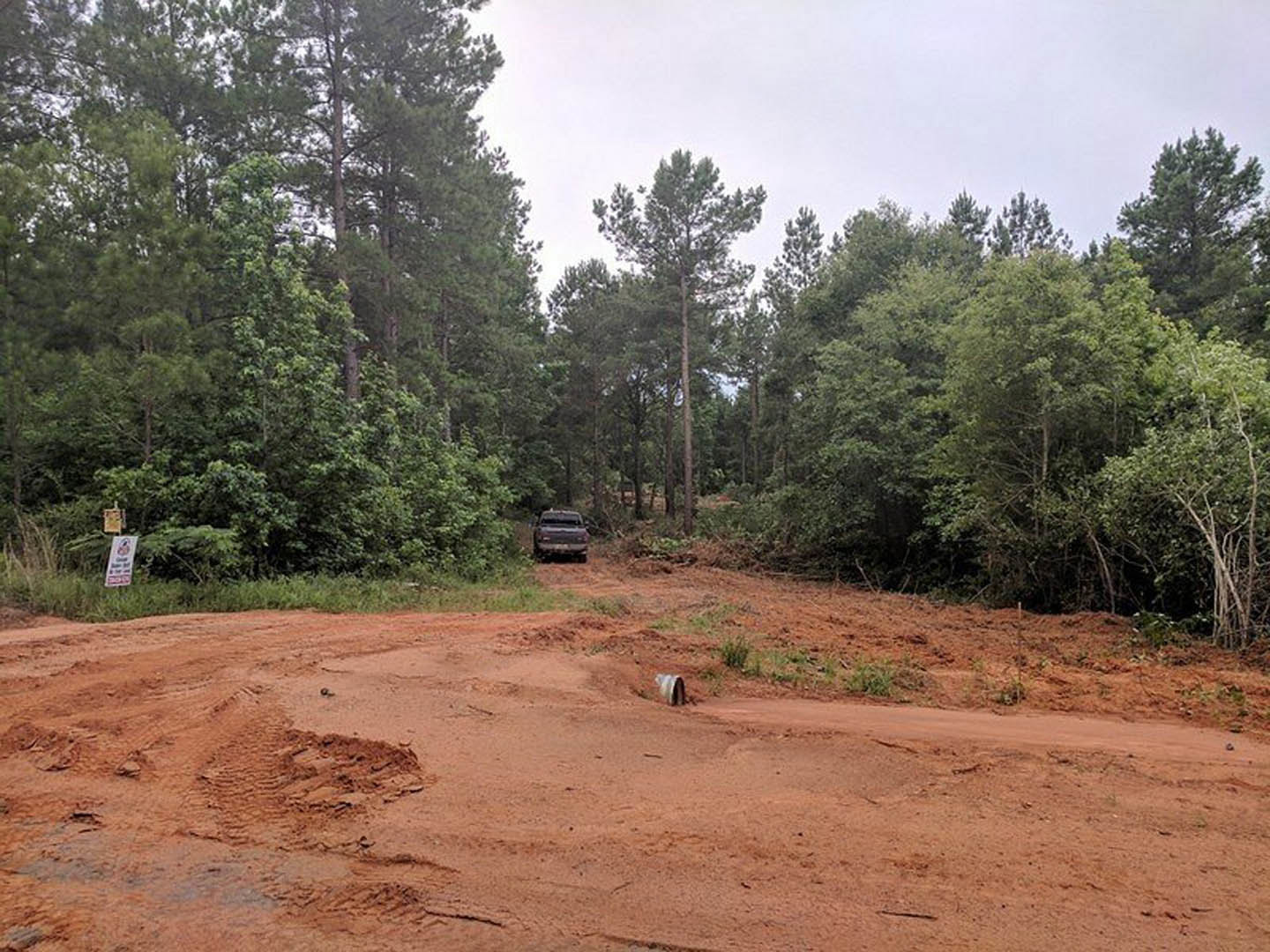 A dirt road winding through dense forest, with a car parked on the soil, surrounded by tall trees and a white sign with black text nearby.