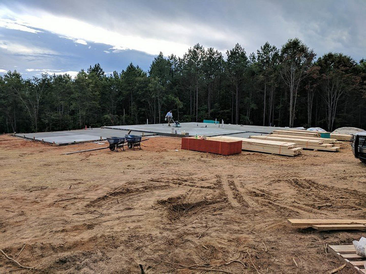 Framed wooden structure on a dirt construction site with wheelbarrows, red brick wall, concrete cutting equipment, and surrounding trees under a cloudy sky