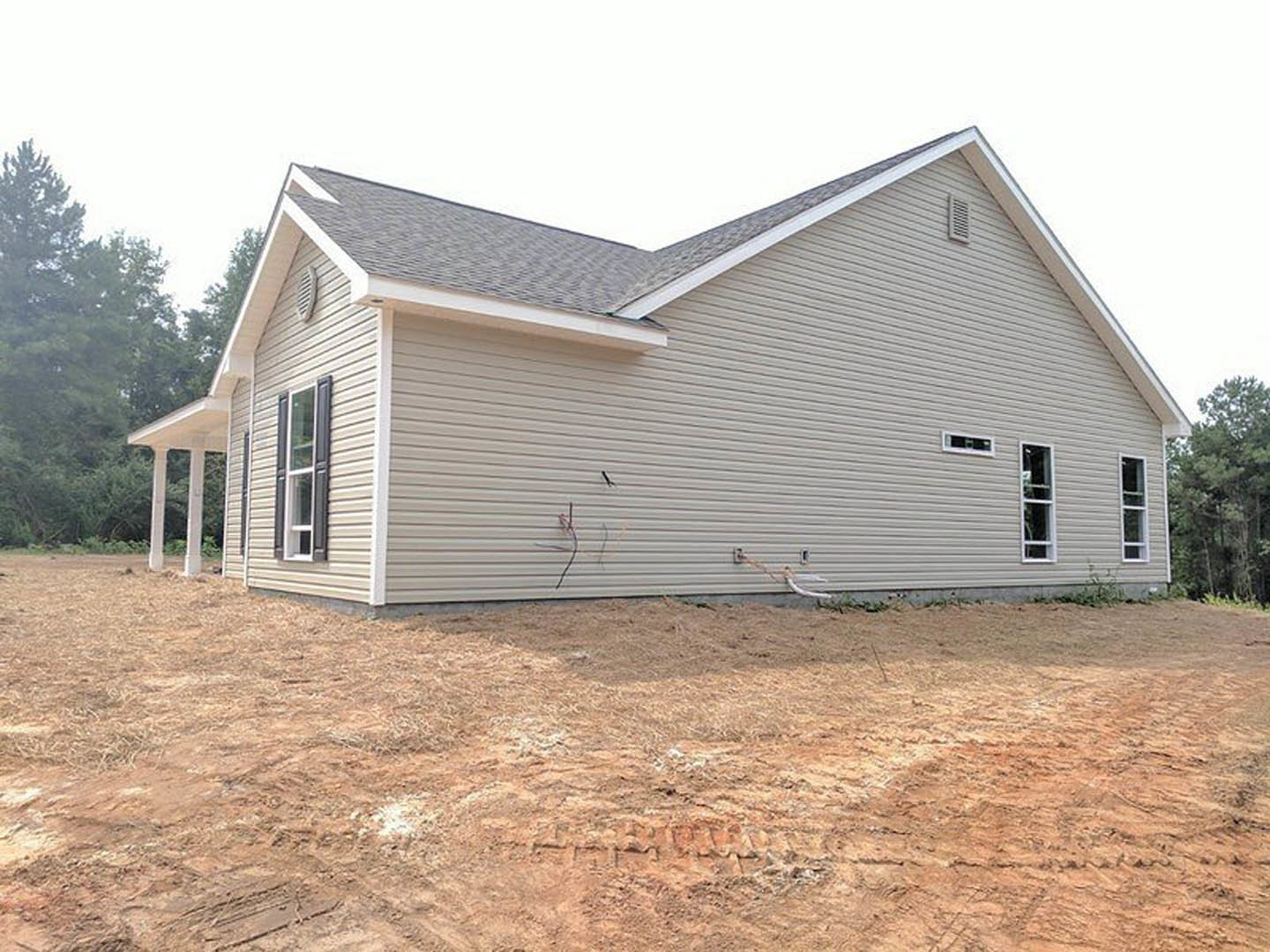 Side view of a house under construction with tire tracks in dirt, white siding, large window, and a leafy tree nearby