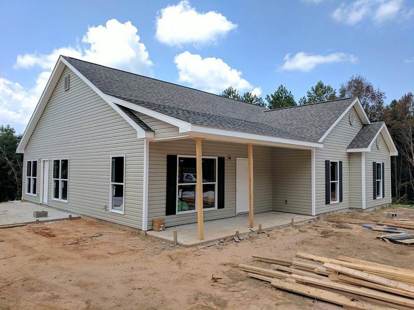 Two-story house under construction with exposed wood framing, unfinished porch, white-framed windows, stack of brown boxes and wood planks on dirt ground, cloudy sky and trees in