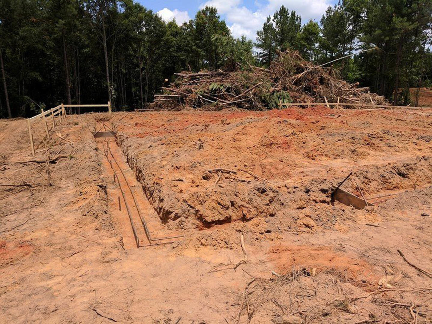 Dirt field with scattered metal rods, trees and bushes in the background, fenced dirt hill, and a long trench under a cloudy sky