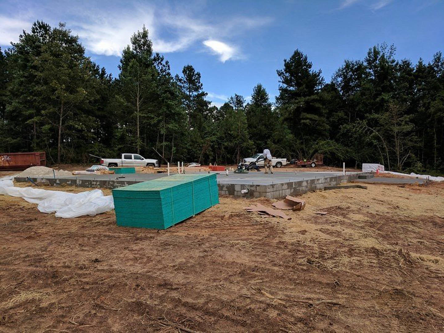 Partially built house framed with wood, blue insulation panels stacked nearby, white truck parked beside construction materials, green dumpster next to dirt pile, scattered debris