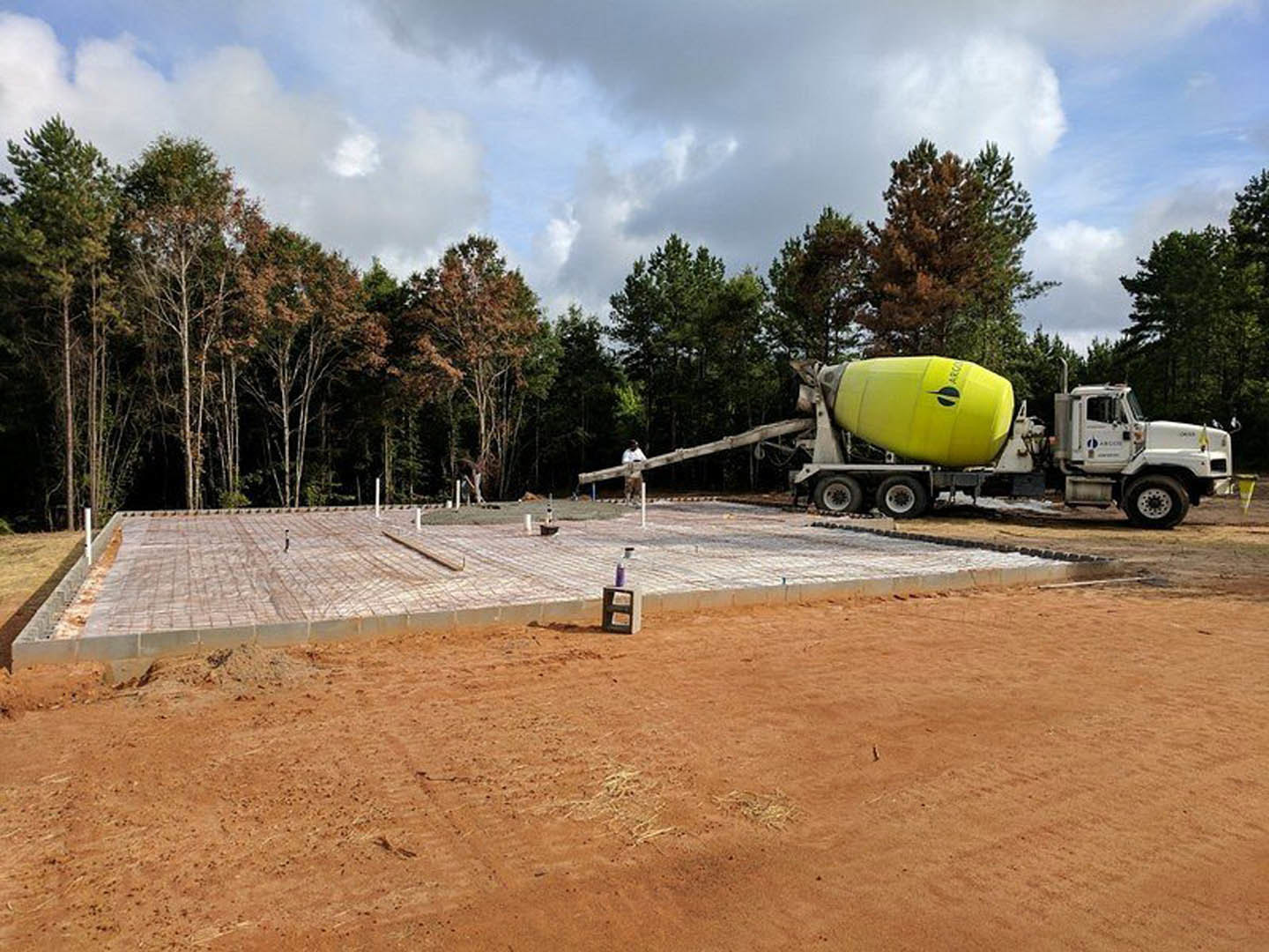 Cement mixer truck parked on dirt construction site beside concrete block and spray can, surrounded by trees under cloudy sky