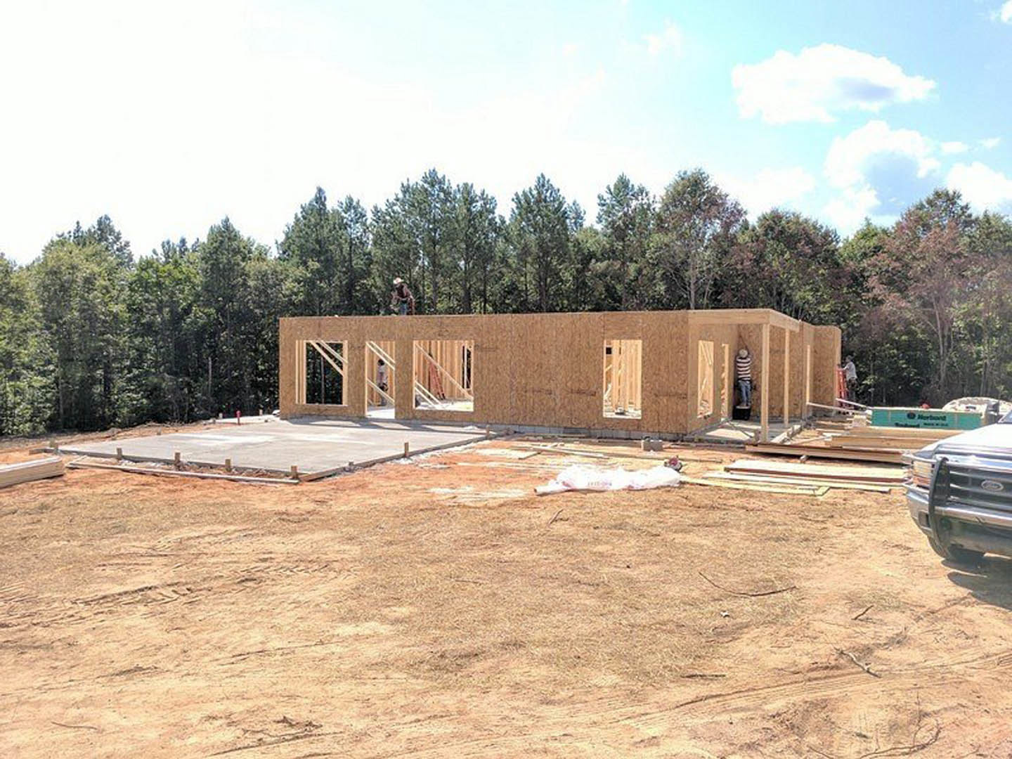 Wood-framed house under construction with exposed beams, concrete driveway, surrounding trees, and blue sky with scattered white clouds