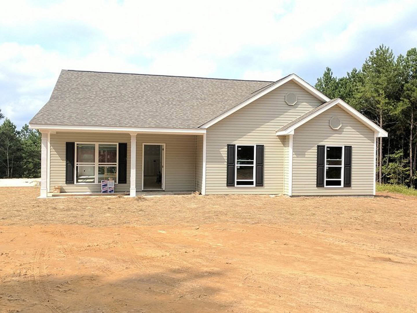 Wood-framed house under construction with exposed porch, open front door, white-trimmed windows, dirt yard, and surrounding trees