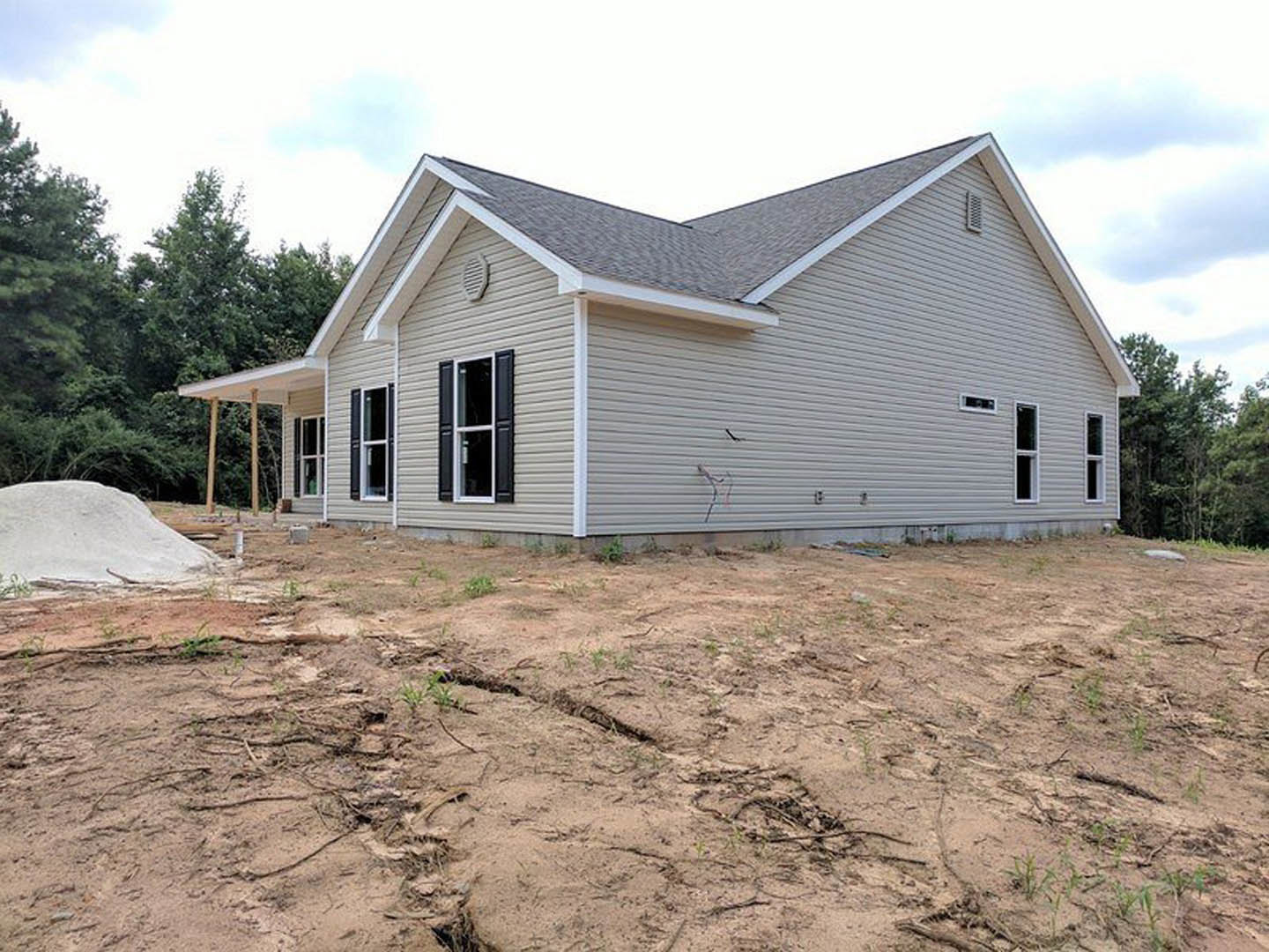 Partially built home with gray siding, white-framed windows, covered porch, exposed dirt yard, large white rock, and cracked ground under cloudy sky.