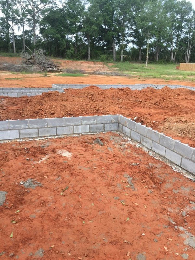 Red dirt foundation bordered by a brick wall, scattered piles of soil and branches, grassy patches, and trees in the background.