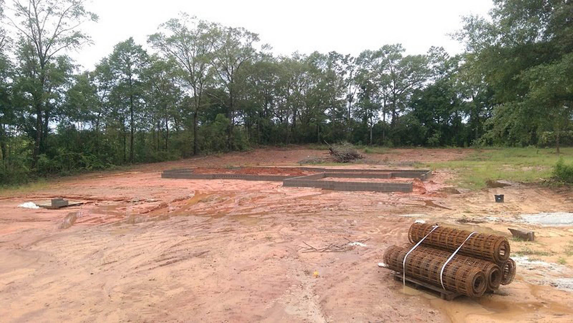 Dirt construction site bordered by a wooden fence, scattered bricks and metal rolls, with tall trees and forested background