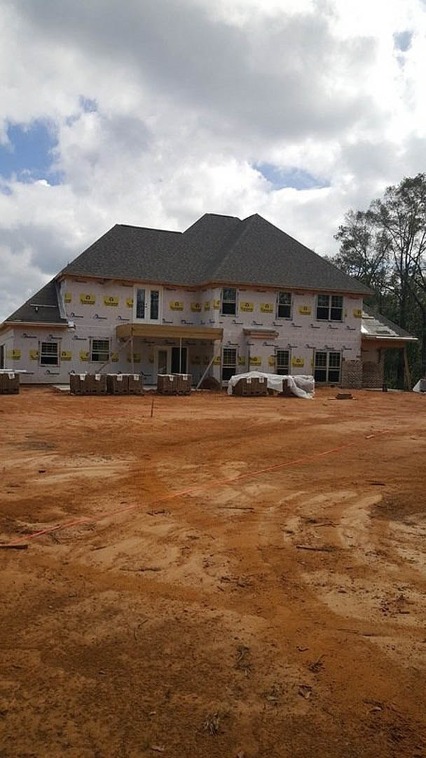 Two-story house under construction with exposed framing, gray roof, and yellow caution signs, surrounded by red dirt lot and trees in the background