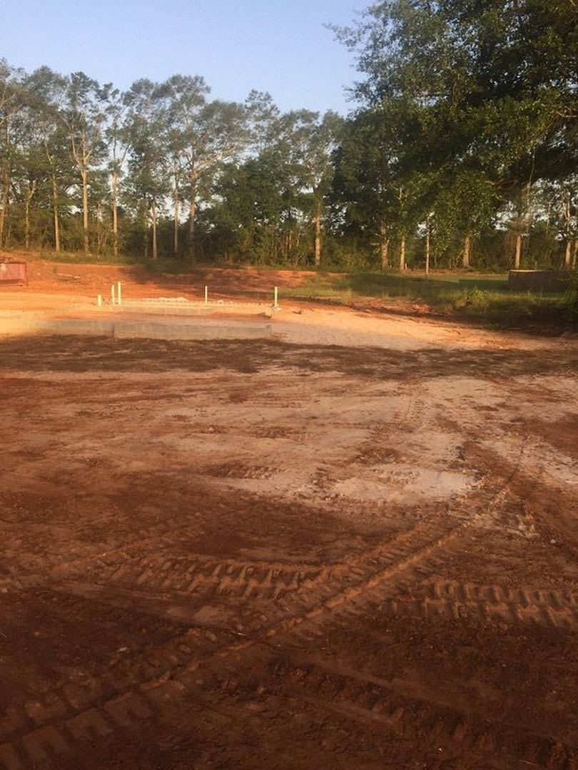 Dirt area with tire tracks bordered by trees under blue sky, blurred building in background