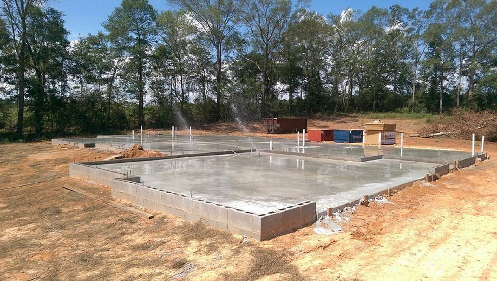 Concrete foundation outdoors with water spraying from embedded sprinklers, surrounded by trees and sky